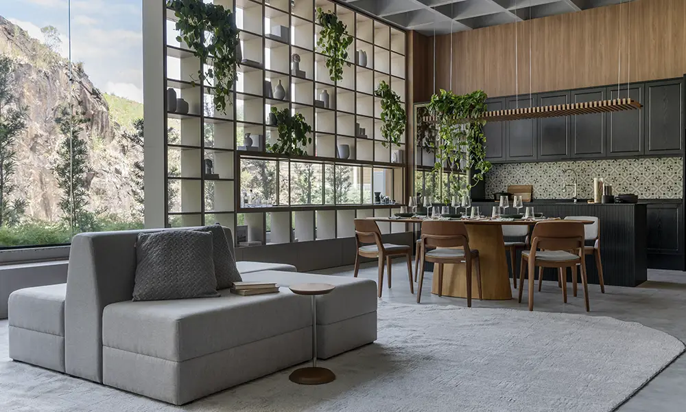 Sala de estar e jantar moderna com mobiliário minimalista, grandes janelas com vista para a natureza, parede com prateleiras preenchidas por plantas e cozinha integrada em tons de madeira e preto.